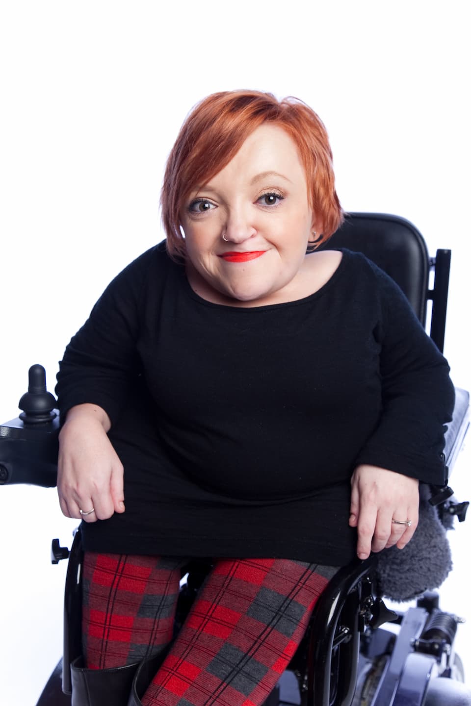 Studio photo of Stella in her wheelchair smiling at the camera. She's wearing a black top and red tartan pants.
