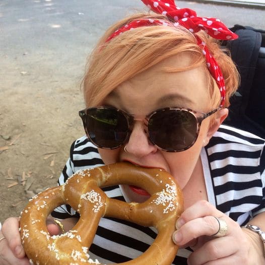 Stella has a polka dotted scarf in her hair and wears a striped T shirt. She is taking a bite from a large pretzel.